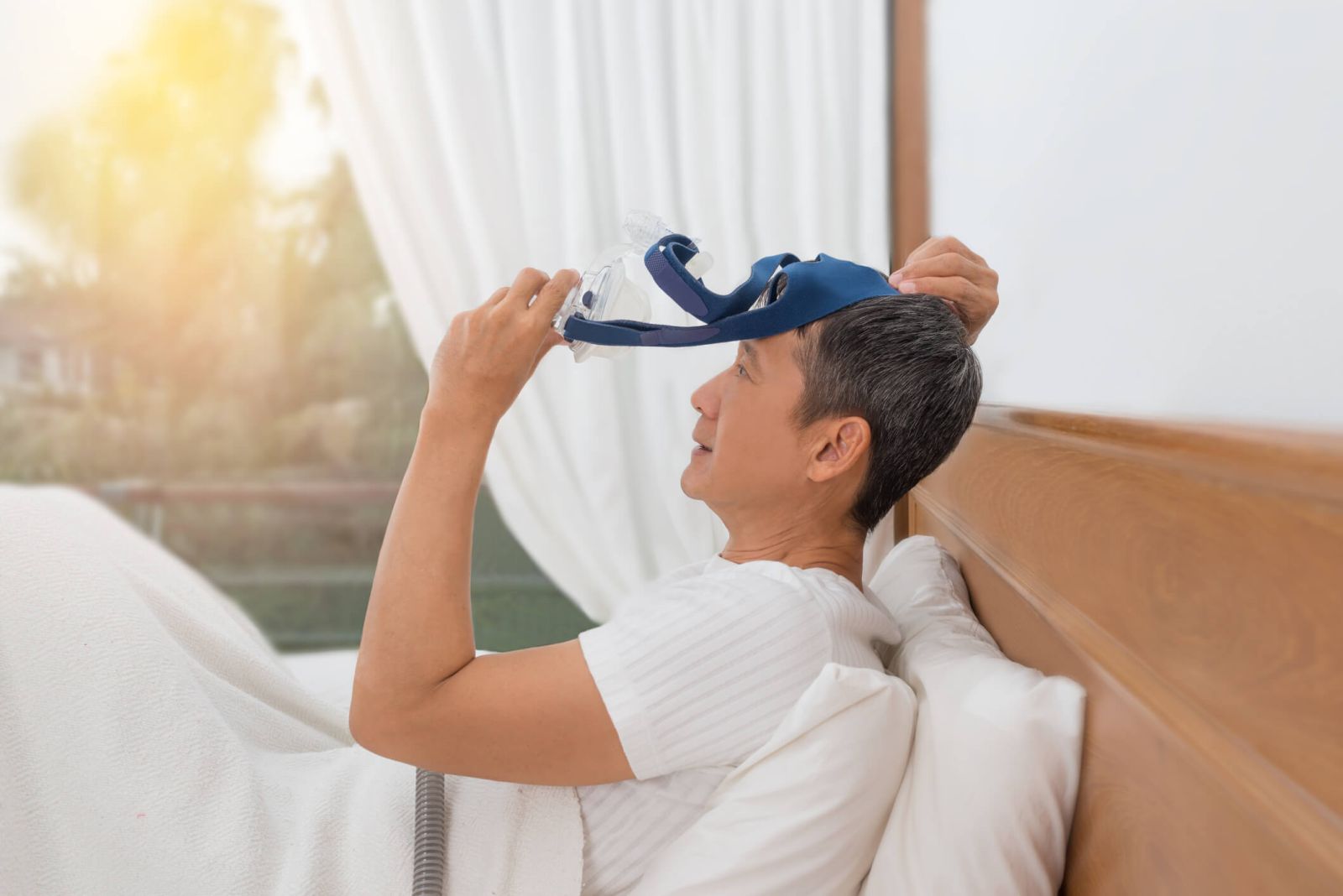 Asian man sitting in bed and putting on a CPAP mask